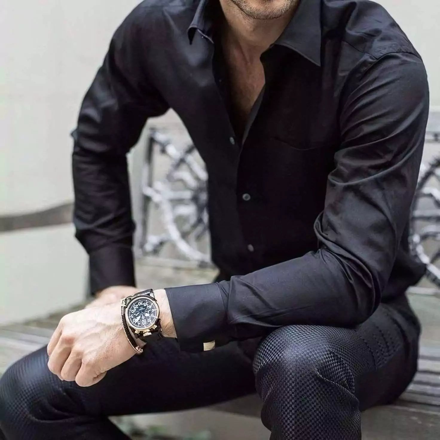 Man wearing a black shirt and dark pants with a watch on his wrist, sitting on a bench.
